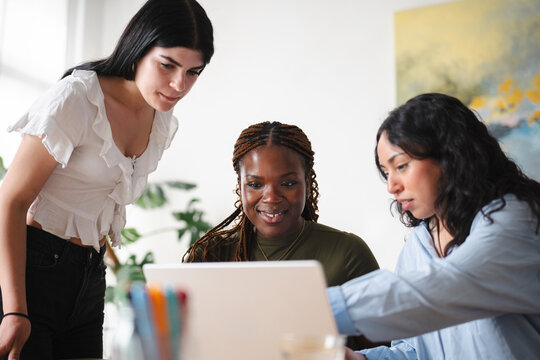 Three diverse female colleagues collaborate in a bright office. They gather around a laptop computer, pointing at the screen and discussing a project together.