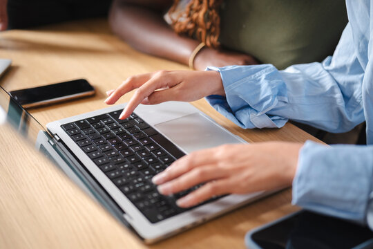 A woman types on a modern laptop at a wooden office desk. Her colleague sits close by as they collaborate on a professional project, focusing on daily tasks together.