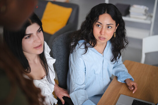 Three young female colleagues collaborate in a bright modern office. Two women sit at a wooden desk with a laptop while listening intently to a teammate during a meeting.
