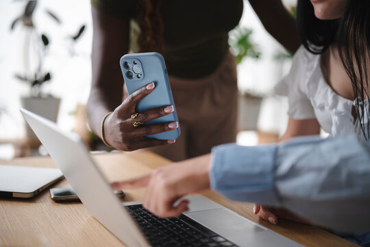 Three female colleagues collaborate in a bright office, gathering around a desk. One holds a smartphone while another points at the laptop screen during a business meeting.