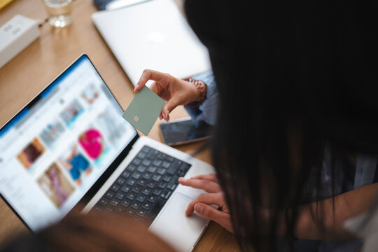 A woman with dark hair sits at a wooden desk, holding a credit card while shopping online with her laptop computer. She is making an electronic payment for clothing.
