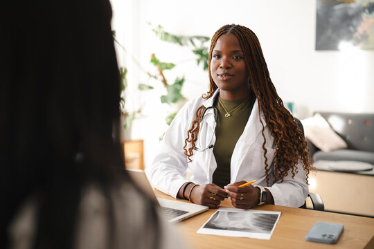 A young female doctor with braided hair sits at her desk and consults with a patient. She holds a pen and reviews medical scans during the appointment in her office.