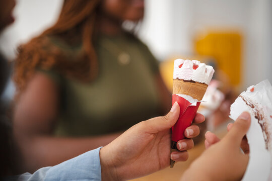 A young woman holds a sweet ice cream cone, peeling back the red paper wrapper. Her friends sit in the blurred background, enjoying a casual afternoon snack together.