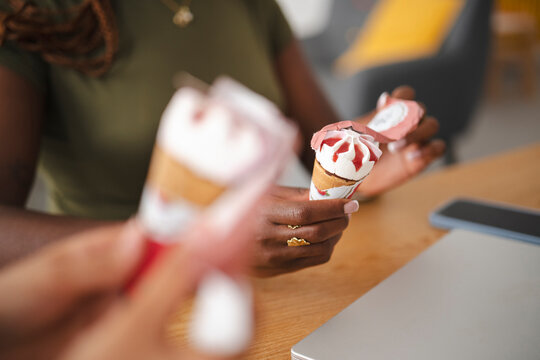 An African American woman sits at a wooden desk and unwraps a delicious ice cream cone with strawberry topping. Her coworker holds another dessert in the foreground.