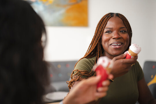 Two young Black women share ice cream cones and smile indoors. The friend in focus holds a treat while talking to her companion in a casual living room setting.