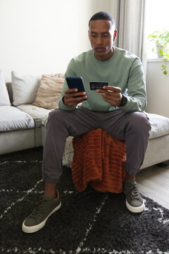 Adult African American male holding phone and tapping credit card sitting on ottoman in living room