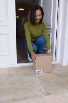 African American woman crouching at front doorway handling cardboard box with barcode label