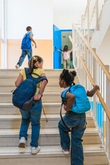 Diverse elementary school children with backpacks walking up stairs in a bright school building, heading to their classrooms © Koldo_Studio