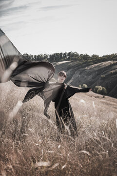 Young woman raises sweeping black textile overhead in dry field with rebellious energy. Defiance, self-expression, creative liberation concept