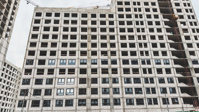 Unfinished multistory residential building with concrete frame, white brick walls, empty window openings, new windows on lower floors, tower crane, concept of housing construction, real estate
