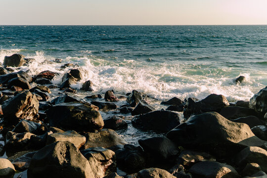 White ocean wave foam crashing against dark volcanic rocks on a rocky shoreline under warm sunlight.   
