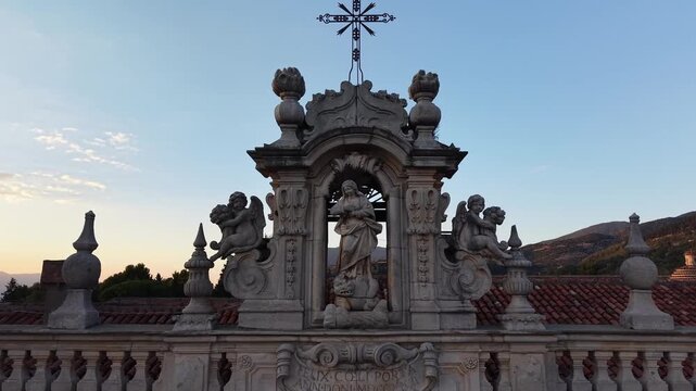 Baroque statue and wrought iron cross crowning the facade of the Certosa di San Lorenzo in Padula at sunset, Campania, Italy