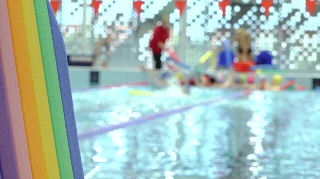 Bright multicolored foam pool noodles frame an indoor swimming pool, with sparkling water, colorful equipment, and decorative lights creating a playful atmosphere and copy space.