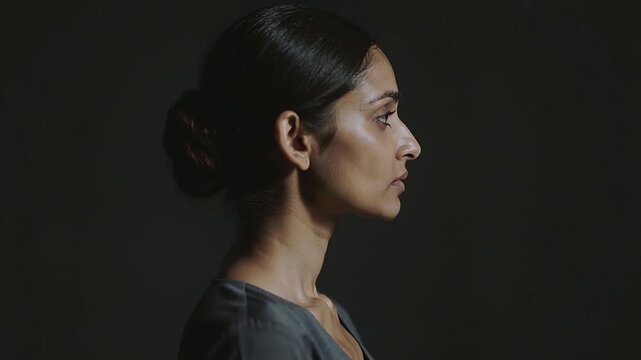 Turning woman rotating head right on pose cue in studio, dark backdrop, grey V-neck, copy space