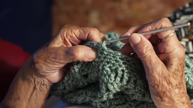 NOA, ITALY - September 23, 2026. Close-up of an elderly woman's weathered hands skillfully crocheting with a hook, working sparkly green yarn into a textured stitch pattern. Shot indoors in warm light