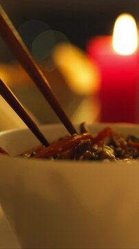 A hand picks food from a bowl with chopsticks at a restaurant dining table. The warm candlelit setting provides soft illumination for the meal, capturing a quiet moment of eating