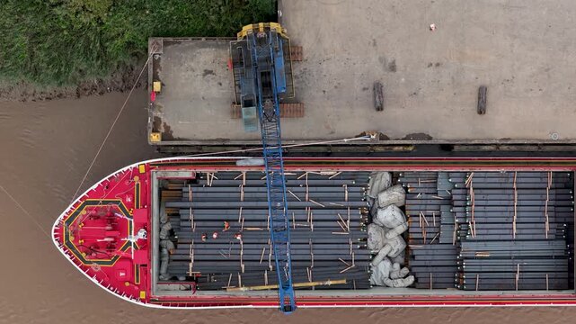 Crane Moving Heavy Metal Pipes off of a Red Cargo Barge at the Dock