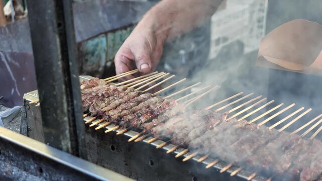 SLOW MOTION: hand of chef turning rows of authentic Italian arrosticini lamb skewers cooking on a smoking charcoal grill, preparing a popular street food from the Abruzzo region in Italy