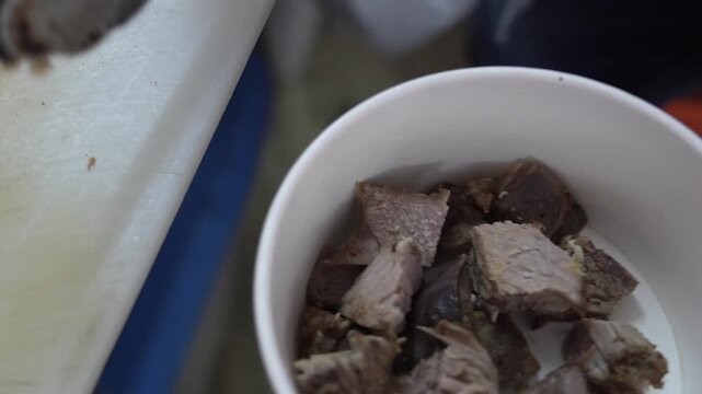 Close-up 4K footage of hand using tongs cutting boiled beef meat chunks on chopping board and putting into white bowl for soup, street food preparation.