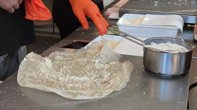 Hand in orange glove folding thin. Crispy borlengo filled with grated Parmigiano Reggiano cheese in Italy. traditional Italian street food from Emilia region being prepared on stainless steel surface