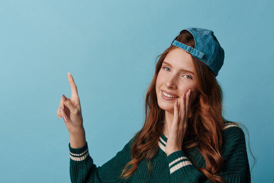 Smiling young woman in a green sweater and backwards blue cap points her finger up while touching her cheek. Studio shot on a blue background. Concept of idea or promotion.
