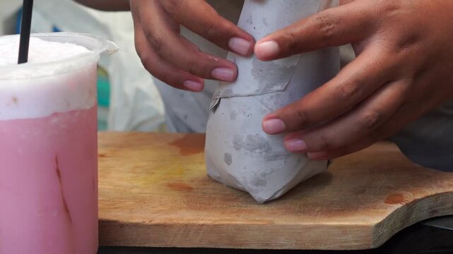 Person untying rubber band and opening traditional brown paper takeaway meal outdoors.