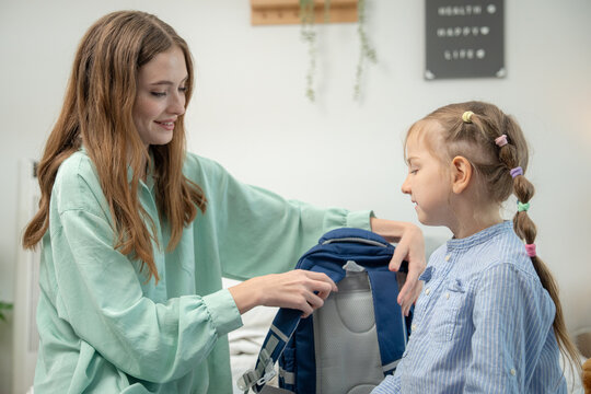 Mother supporting daughter wearing backpack while holding teddy bear on bed. Young mother smiling and supporting cute schoolgirl carrying backpack and stuffed toy bear in bedroom.