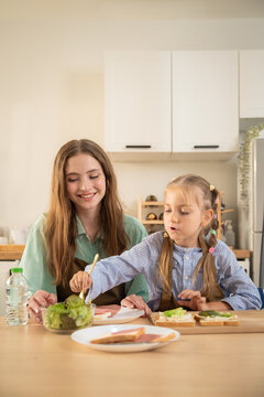 Happy little girl talking excitedly to mother while sitting at kitchen table with food. Cheerful daughter sharing funny moment with smiling mother during healthy meal preparation in home kitchen.