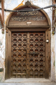 Arched old wooden door with Arabic calligraphy above and square panels with iron studs on a weathered whitewashed wall with a light switch and cables, Stone Town, Tanzania