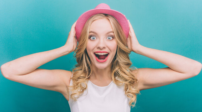 Very exited girl is looking straight forward and holding her pink hat with hands on a head. She is surprised a lot. Isolated on blue background.
