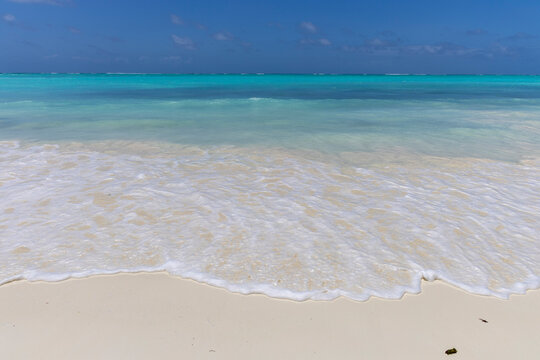 Foamy wave washing over smooth white sand on a tropical beach with turquoise water stretching to the horizon under a deep blue sky with scattered small clouds, Nungwi, Tanzania