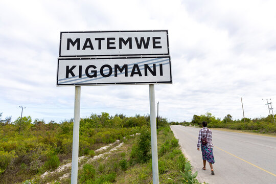 Woman with braided hair and a small bag walking along a road past a Matemwe Kigomani village boundary sign under a cloudy sky with green roadside bushes, Matemwe, Tanzania