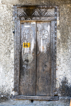Weathered carved wooden double door with floral rosette panels and an ornate frame set in a crumbling whitewashed coral stone wall marked with the number 34, Jambiani, Tanzania