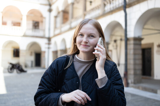 Half-length portrait of a happy caucasian woman calling on a mobile phone, walking outdoors in front of a beautiful historic building with classic arches and columns. 