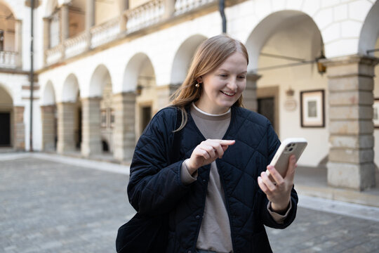 Half-length portrait of a happy caucasian woman looking at a mobile phone screen while walking outdoors in front of a historic building with classic stone arches. 
