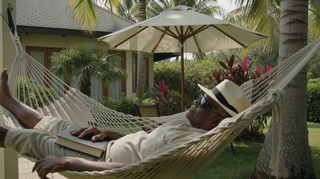 Hammock shifting, lying senior in straw hat, shades tilting head, resting on closed book in garden