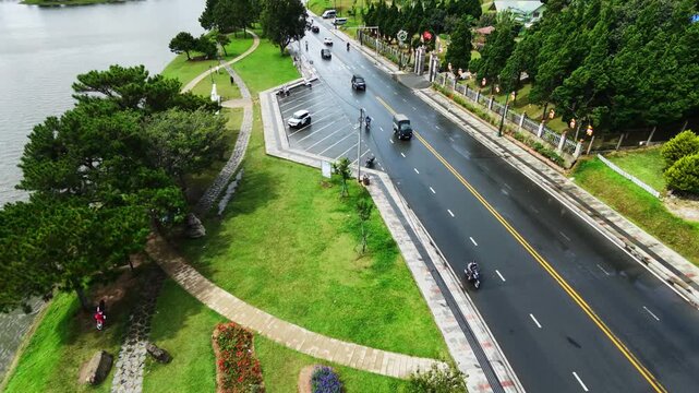 Aerial tracking shot of vehicles moving along treelined riverside road in daylight