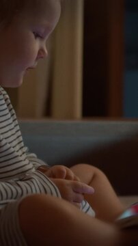 A young child sits cozily on a sofa inside a home living room, holding a smartphone and watching cartoons, framed by family childcare and parenting routines involving modern technology devices