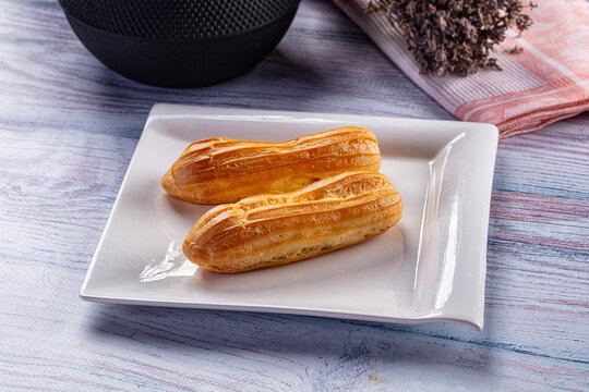 Two golden brown eclair pastries on white square plate with textured wood surface and dried lavender bundle nearby as dessert