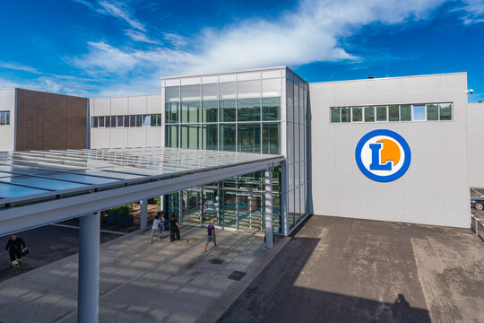Exterior of an E.Leclerc hypermarket in Incarville, Eure, Normandy, showing the large blue lettering, the circular logo and the glass entrance under a bright blue sky.