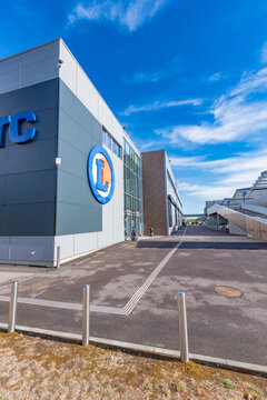 Exterior of an E.Leclerc hypermarket in Incarville, Eure, Normandy, showing the large blue lettering, the circular logo and the glass entrance under a bright blue sky.