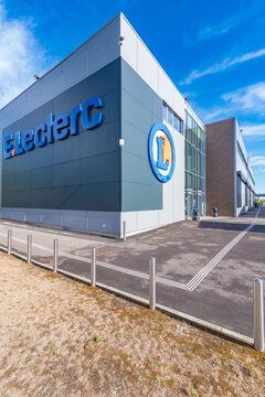 Exterior of an E.Leclerc hypermarket in Incarville, Eure, Normandy, showing the large blue lettering, the circular logo and the glass entrance under a bright blue sky.