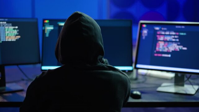 Back view of a hooded hacker typing code on multiple illuminated computer monitors in a dark room, representing cybersecurity threats, data breaches, cyber attacks, and advanced software programming.