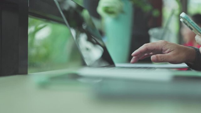 Freelance worker typing on keyboard during online tasks, typing messages beside phone as remote typing continues beside window.