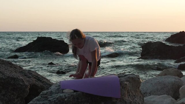 Athletic child executing advanced balancing pose on beach boulders. Static wide shot displays determined gymnast training beside rolling waves during golden hour.