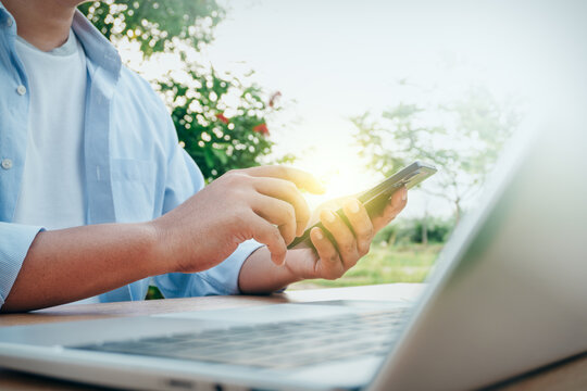 Man using smartphone and laptop computer at a wooden table in an outdoor green nature setting, concept of mobile technology, remote work, and freelance lifestyle.