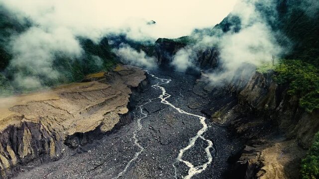 Aerial footage travels above a braided river cutting through a rugged mountain canyon as dense mist moves between steep green slopes. The dramatic landscape highlights erosion, freshwater processes, g