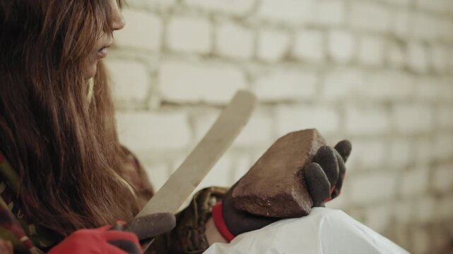 Close up of orphan sharpening cutlass blade on stone with gloved hand, face lowered in concentration as sharp metal edge presses firmly against rough block