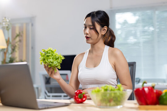 Asian woman discussing healthy nutrition and diet online. Young woman video calling on laptop, holding lettuce while promoting healthy eating and balanced lifestyle.