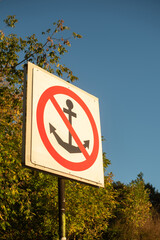A no anchoring sign stands against a clear blue sky. The sign features a red circle with a diagonal line over an anchor symbol. Surrounding trees add greenery. © Ksu